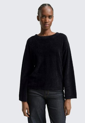 Pull noir, moelleux à manches longues avec un décolleté large et une coupe décontractée, associé à un jean foncé. Texture douce et design simple.