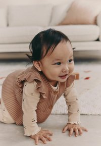 Niño en un chaleco marrón acanalado con volantes y botones, vistiendo una camiseta de manga larga color crema con un patrón floral, gateando sobre una superficie clara.