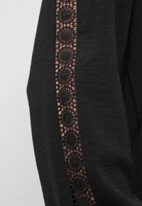 Μαύρο ύφασμα με υφή επιφάνειας που διαθέτει δαντελωτό πάνελ διακοσμημένο με κυκλικά ανθισμένα σχέδια κατά μήκος μιας πλευράς.