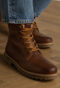 Braune Lederstiefeletten mit runder Zehenpartie, Schnürverschluss, Gummisohle und kontrastierenden beigen Schnürsenkeln. Strukturierte Oberfläche mit Markenlogo an der Seite.