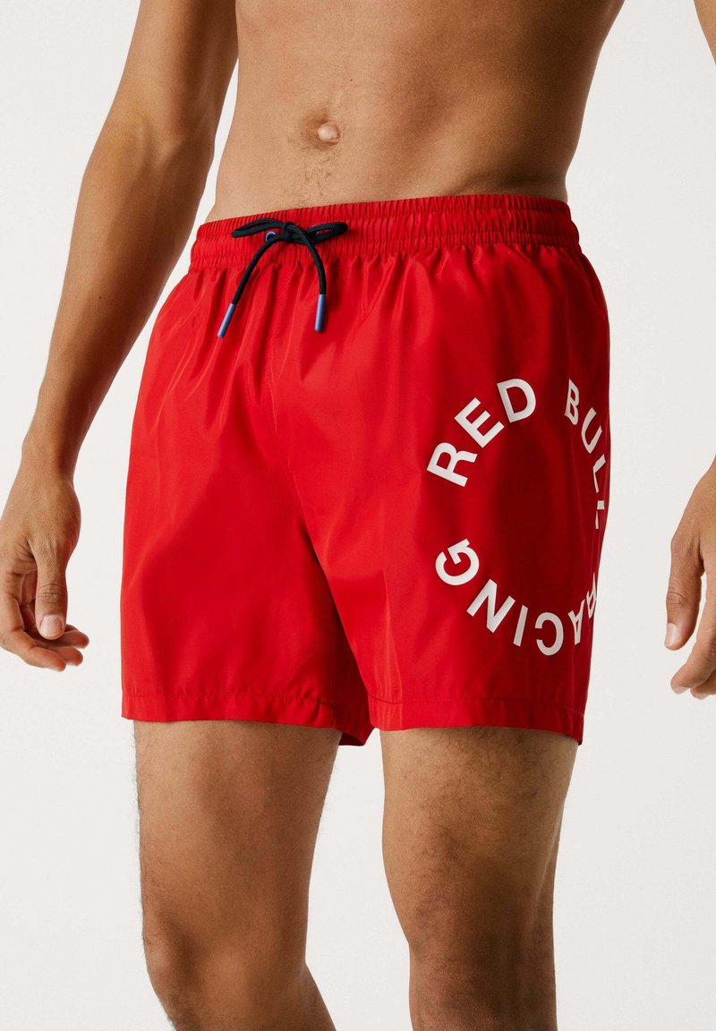 Bañadores rojos de tela ligera, con cinturilla ajustable mediante cordón y un audaz texto en blanco "RED BULL" en un patrón circular en la pierna.