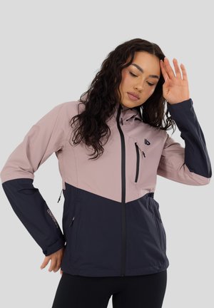 Frau richtet Haare und trägt eine rosa-navy Reißverschlussjacke mit Brust- und Seitentaschen vor hellem grauem Hintergrund.