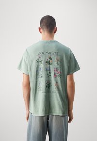 Muž v bledě zeleném tričku s devíti botanickými květinovými ilustracemi a nápisy "BOTANICALS" a "KEEP THINGS WILD" na zadní straně.