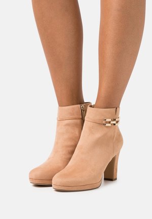 Bottines beiges en suède avec talons carrés, fermetures éclair latérales et détail de chaîne dorée portées sur des jambes nues sur fond blanc.