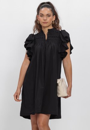 Vrouw met lang haar in een paardenstaart, die een zwarte mouwloze jurk met ruches draagt en een lichtbeige handtas vasthoudt tegen een effen achtergrond.