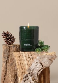 Green glass candle with a geometric pattern, lit flame, resting on a wood surface alongside a pinecone and sprigs of greenery.