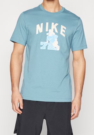 Homme portant un t-shirt Nike bleu clair avec un personnage de chien de dessin animé et le numéro 72, associé à un short noir, torse visible.