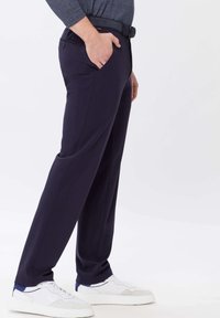 Homme portant un pantalon bleu marine avec la main dans la poche, associé à des baskets blanches sur fond clair.