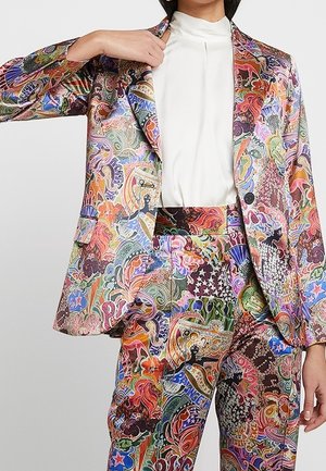 Persona con un traje multicolor de estampado abstracto y una blusa blanca de cuello alto, ajustándose el cuello de la chaqueta con una mano.