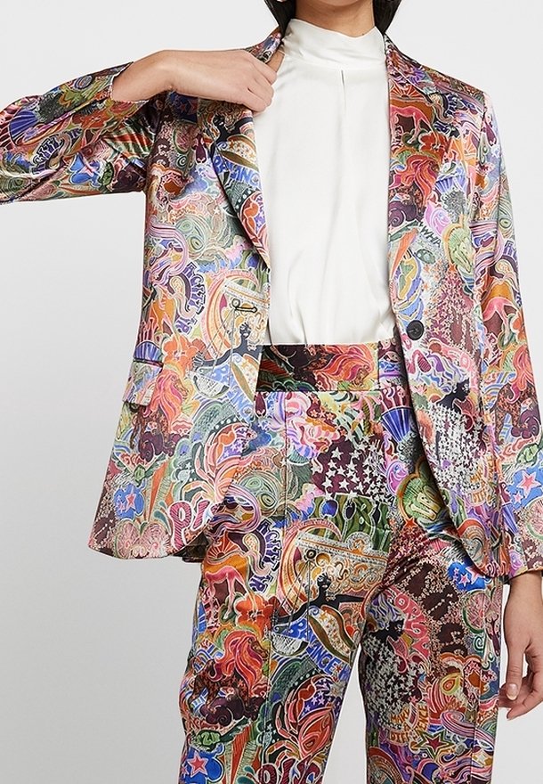 Personne portant un costume multicolore à motifs abstraits avec un chemisier blanc à col montant, ajustant le col de la veste d'une main.