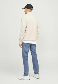 Man met een zwarte pet, beige sweater, wit overhemd, blauwe spijkerbroek en witte sneakers, staand met de rug naar voren op een eenvoudige lichte achtergrond.