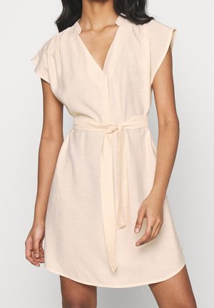 Woman wearing a sleeveless, light beige dress with V-neck and fabric belt tied at the waist, standing against a plain background.