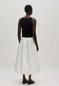 Schwarzes ärmelloses Strickoberteil, kombiniert mit einem weißen, voluminösen Rock. Der Rock hat eine glatte Oberfläche und einen elastischen Bund. Schwarze Schuhe runden das Outfit ab.