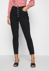 Woman wearing high-waist black skinny jeans with button fly, paired with black open-toe heeled sandals and a red floral long-sleeve top.