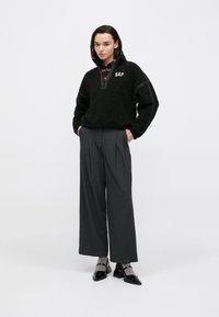 Maglione in pile nero con cerniera e logo "GAP", abbinato a pantaloni ampi grigi. Il modello è in piedi con le mani nelle tasche, indossa scarpe nere.