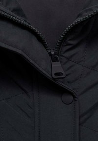 Section de veste noire matelassée avec une fermeture éclair et un bouton-pression. Présente une texture lisse et mate ainsi qu'un col haut. La fermeture éclair se trouve sur le côté gauche.