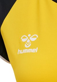 Gul atletisk skjorte med sorte accenter; har glat stof, Hummel-logo i hvid, og et geometrisk mønster på forsiden.