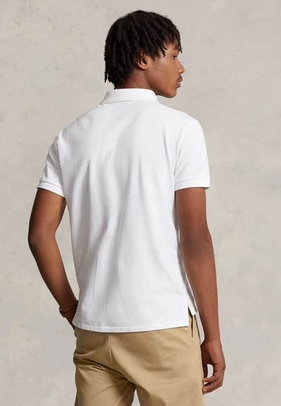 Jeune homme avec de courtes dreadlocks portant un polo blanc uni à manches courtes et un pantalon beige, vu de dos.
