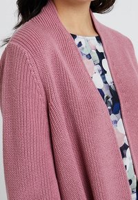 Cardigan rosa lavorato a maglia con un motivo texture, aperto davanti e colletto a costine, indossato sopra una blusa multicolore con motivi circolari.