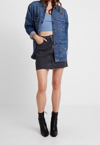 Giacca di jeans con chiusura a bottone, cinque tasche e ricamo verde, abbinata a una gonna di jeans grigia con orli sfrangiati e stivali neri alla caviglia.