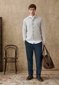 Graue strukturierte Strickjacke mit Knopfleiste, darunter ein weißes Hemd, dunkelblaue Hose, braune Leder Schuhe und braune Wildledertasche. Holzstuhl im Hintergrund.