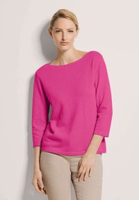 Frau mit blonden Haaren, die ein leuchtend pinkes Langarmoberteil und beige Hosen trägt, steht vor einer hellen Wand, schaut geradeaus mit einem neutralen Ausdruck.