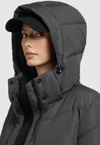 Black puffer jacket with a hood, featuring a zip closure, snap buttons, and stitching details. Worn with a black cap. Soft texture.