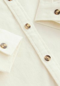 Chemise boutonnée couleur crème avec un tissu texturé, deux poches poitrine et des boutons ronds sombres avec un léger motif dégradé.