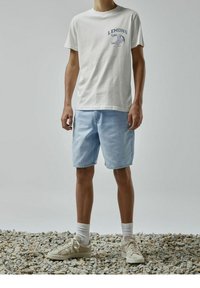 Camiseta blanca de algodón con gráfico azul "LEMONS" en el pecho, combinada con pantalones cortos de mezclilla azul claro y zapatillas beige con calcetines blancos.