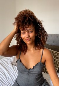 Harmaa ribbitekstuurinen toppi röyhelöreunuksilla, jossa on syvä pääntie. Mallilla on runsaat kiharat hiukset, ja hän istuu valkoisella vuodevaatteella vuodatulla sängyllä.