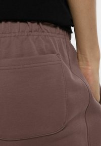Close-up van een persoon in mauve elastische taillebroek met een achterkantzak en een gedeeltelijk zichtbare zwarte top.