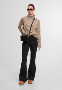 Beige Rollkragenpullover, schwarze weit geschnittene Jeans und braune Plateauschuhe. Kleine schwarze Umhängetasche mit Reißverschluss. Transparente Sonnenbrille.