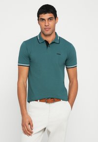 Türkizkék galléros póló fekete gallérral, fehér és tengerészkék csíkozású ujjakkal, valamint egy kis fekete logóval a mellrészen. Világos színű nadrággal viselve.