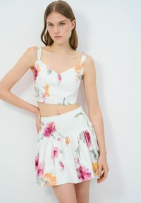 Weißes Blumen-Crop-Top mit dünnen Trägern, kombiniert mit einem passenden, ausgestellten Rock. Merkmale sind Aquarellmuster in Pink, Orange und Grün.