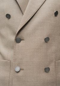 Μπεζ υφασμάτινο σακάκι διπλής σειράς με έξι στρογγυλά μεταλλικά κουμπιά και δύο εμπρόσθιες τσέπες.