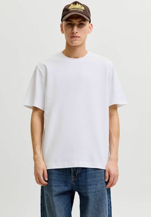 Junger Mann trägt ein schlichtes weißes oversized T-Shirt, blaue Jeans und eine braune Baseballkappe mit gelber Stadtsilhouetten-Stickerei.