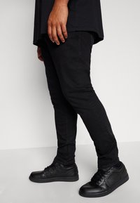 Svarta denimjeans med slim fit, parat med svarta lågtoppsneakers. Outfiten betonar ett monokromatiskt färgschema.