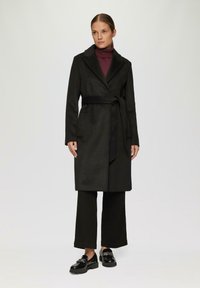 Zwart wollen blend wikkeljas met een sjaalkraag, zijzakken, gecombineerd met wijde zwarte broeken en glanzende zwarte loafers.