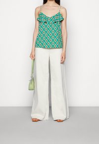Femme portant un pantalon large blanc et un haut cami à motifs verts, tenant un petit sac à main vert clair, se tenant devant un fond blanc uni.