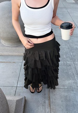 Personne portant un haut blanc sans manches et une jupe noire superposée, tenant un gobelet de café à emporter, debout sur un trottoir carrelé en plein air.