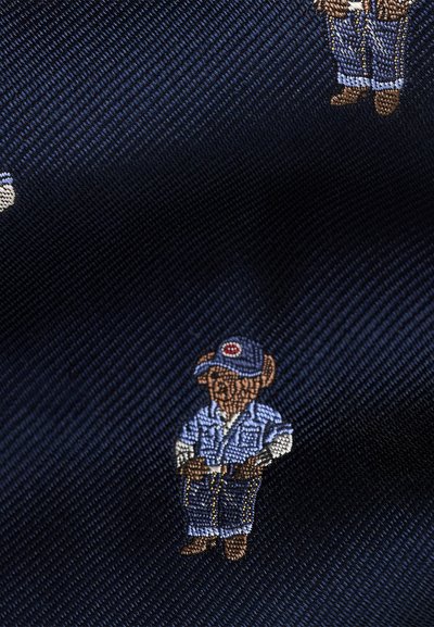 Μικρή κεντημένη αρκούδα που φοράει μπλε καπέλο, ανοιχτό μπλε πουκάμισο και σκούρο μπλε παντελόνι, σε φόντο υφασμάτινης υφής σκούρου μπλε χρώματος.