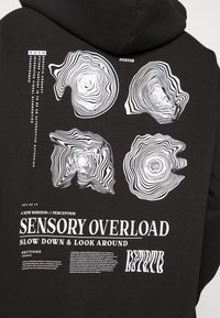 Felpa nera con cappuccio, caratterizzata da una stampa grafica bianca con motivi astratti che ricordano gli anelli degli alberi e testo, tra cui "SENSORY OVERLOAD" e "RUSH."