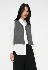 Young woman with straight black hair wearing white long-sleeve shirt, gray pinstriped vest with buttons, and black wide-leg pants.