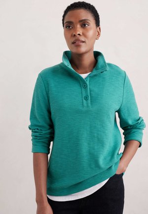 Pull en maille teal avec col montant, deux boutons et poignets côtelés. Tissu léger avec une finition texturée, superposé sur une chemise blanche.