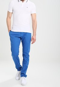 Bílá polo tričko s krátkým rukávem a černým lemem, spárované s modrými slim-fit kalhotami a bílými teniskami. Jednoduchý, čistý design s klasickými barvami.