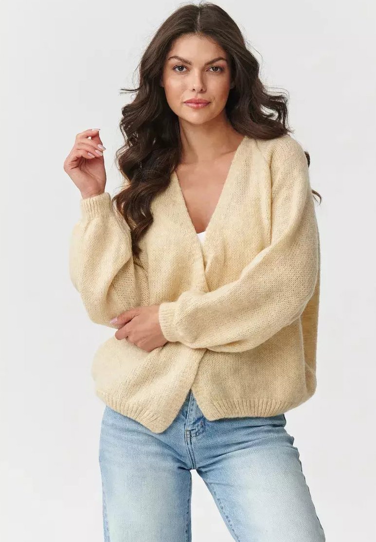 Kremowy, gruby sweter z splotem, z głębokim dekoltem w serek, luźnymi rękawami, teksturowanym materiałem, zapinany z przodu, połączony z jasnoniebieskimi dżinsami.