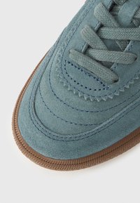 Κοντινό πλάνο μύτης αθλητικού παπουτσιού από σουέντ σε τιρκουάζ χρώμα με λεπτομερείς ραφές, κορδόνια στο ίδιο χρώμα και καφέ λαστιχένια σόλα σε λευκό φόντο.