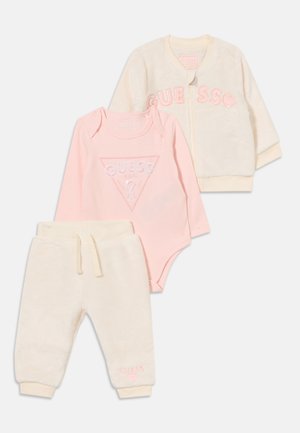 Ensemble bébé doux trois pièces avec pantalon en polaire crème, veste zippée crème et body rose à manches longues, tous siglés Guess.