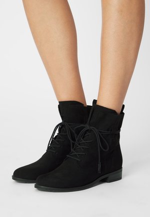 Bottines en daim noir à lacets, portées sur des jambes nues, avec des talons bas et des bouts ronds, sur un fond blanc uni.