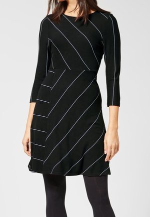 Frau trägt ein schwarzes Kleid mit hellen diagonalen Streifen und Dreiviertelärmeln, kombiniert mit schwarzen Strumpfhosen.